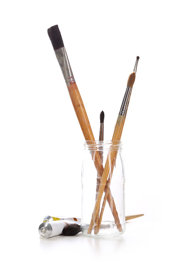 Group of paintbrushes in a glass jar and tubes of paint on a white background.