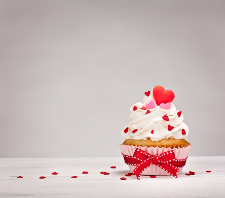 Vanilla Cupcake with buttercream icing, heart shaped sprinkles and a red bow.