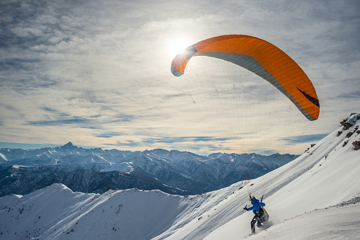 Giaveno, Italy - February 8, 2015: Paraglider running on snowy slope for take off with bright orange kite. Stunning background of the italian Alps in winter season. Shot taken in backlight.