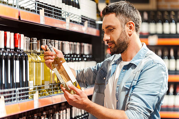 This should be fine. Side view of handsome young man holding bottle of wine and looking at it while standing in a wine store alcohol shop stock pictures, royalty-free photos & images