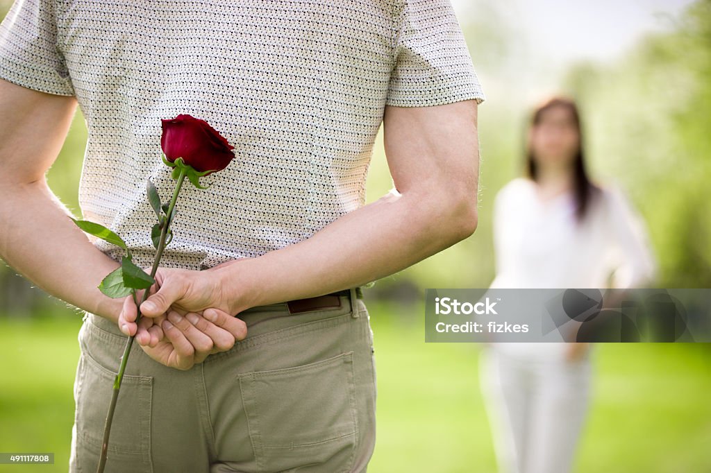 Dating concept Couple of lovers on a date, focus on young man back who is holding red rose while watching his girlfriend approaching Rear View Stock Photo