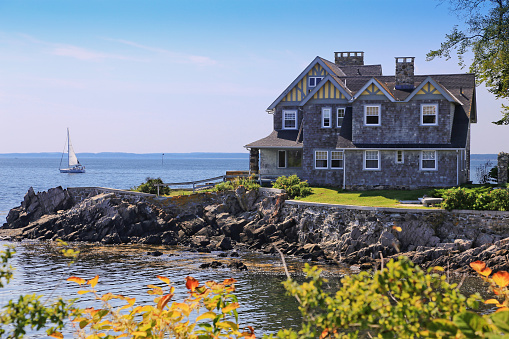 Kennebunkport, ME, USA - September 16, 2015: Luxury Waterfront House with grey shingle exterior, Kennebunkport, Maine, New England, USA. Rocky shore, ocean waters, sailboat, green and yellow bushes, trees, and blue sky with clouds are in the image. Canon EOS 6D (full frame sensor) and Canon EF 24-105mm f/4 L IS lens. HDR photorealistic image.