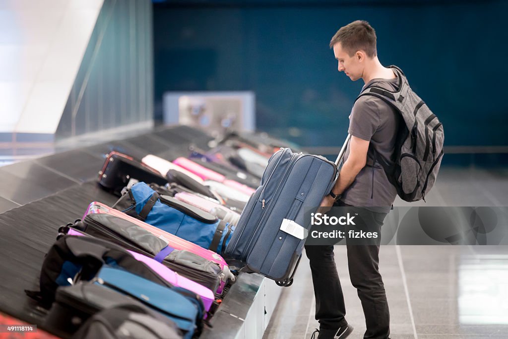 Young man collecting his luggage Young handsome man passenger in 20s with carry-on backpack collecting his luggage at conveyor belt in arrivals lounge of airport terminal building Airport Stock Photo