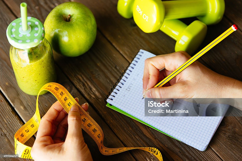 Fitness diet and nutrition routine concept with green detox smoo Woman writing nutrition diet and fitness workout routine. Healthy green detox, apple and dumbbells for slimming down concept. Female hands with measuring tape for checking weight loss. Measuring Stock Photo
