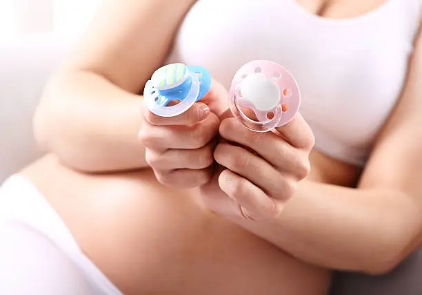 A pregnant woman is holding in his hand the two children's pacifiers blue and pink.