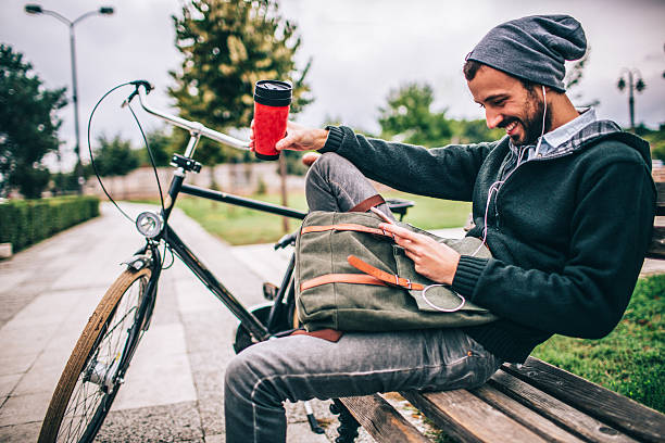 Morning coffee in a park Young man enjoys his hot drink on a bench in a park, while using a mobile phone. His bicycle is parked right besides him. hipster bike stock pictures, royalty-free photos & images