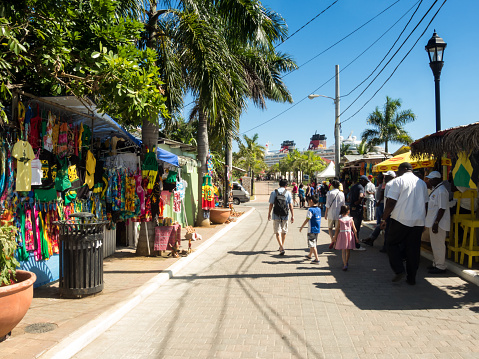 Falmouth, Jamaica – February 12, 2015: View of a street near the main entrance gate at the Falmouth Cruise Port, along with local vendors, tourists, trees and a cruise ship in the background in Falmouth, Jamaica