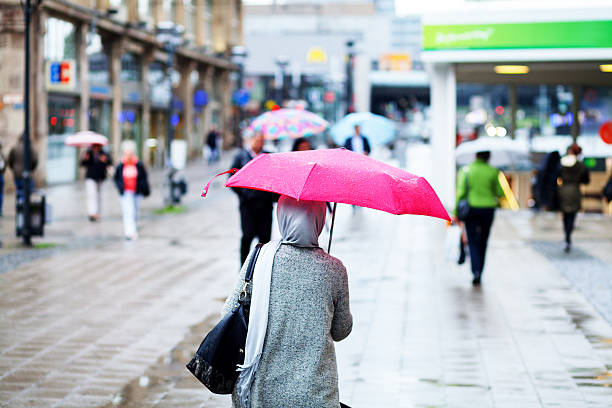Essen, Germany - September 15, 2015: A muslim woman with bag and headscarf is walking with pink umbrella in pedestrian zone Kettwiger Straße in Essen. In background are more people.