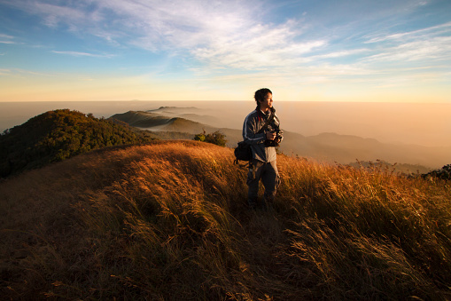 Traveler holding the camera walking in the meadow on top of mountain that around with beautiful view of morning sunrise and blue sky.