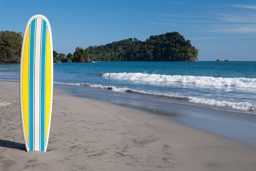 Yellow , blue and white surfboard (off center) on a beach in Costa Rica. Part of Manuel Antonio Rainforest in background.