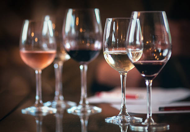 Closeup lima gelas anggur diatur satu di sebelah yang lain, setengah penuh dengan beberapa jenis anggur, anggur merah, putih dan mawar. Kacamata ada di atas meja, ada beberapa kertas dan pena di latar belakang karena ini adalah detail dari winetasting. Menyala dari kedua sisi.