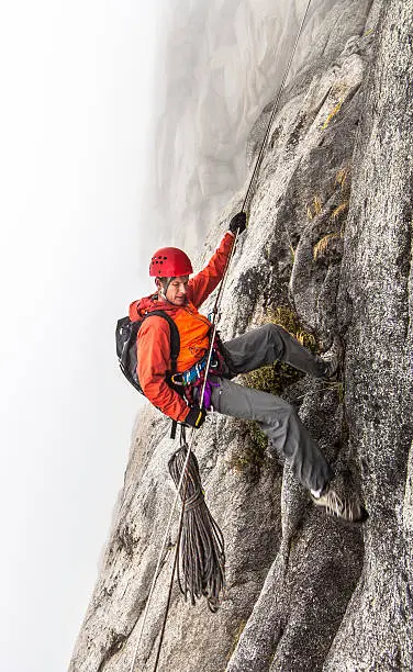 Rock climber wearing a helmet rappels in the clouds during a wet storm on a challenging cliff.