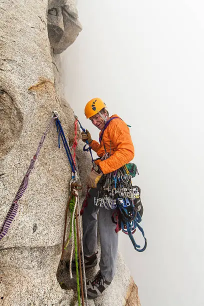 Rock climber wearing a helmet and dangling equipment rappels in the clouds during a wet storm on a challenging cliff.