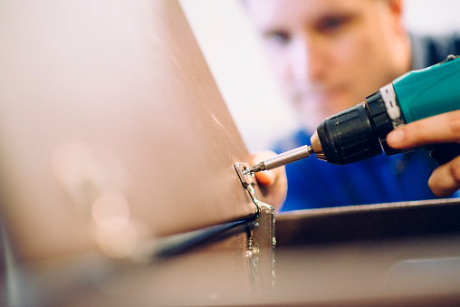 Close-up of carpenter assembling furniture. He is screwing a screw with an electric drill. Selective focus.