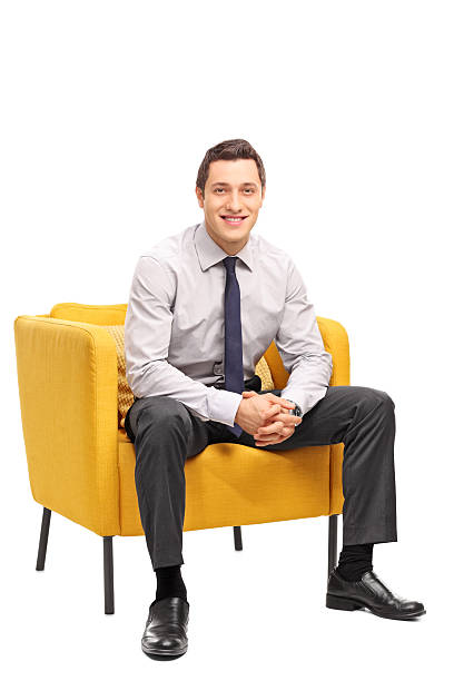 Confident businessman sitting in an armchair Vertical shot of a confident young businessman sitting in a yellow armchair and looking at the camera isolated on white background chairperson stock pictures, royalty-free photos & images