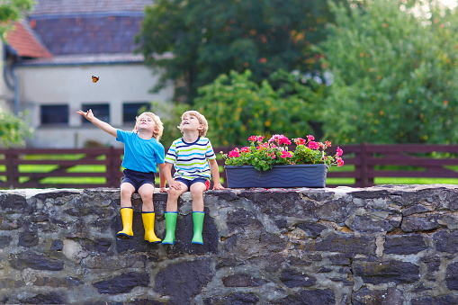 Two little kid boys sitting together on stone bridge in european village. Happy family of two. Twins in colorful gumboots, outdoors.