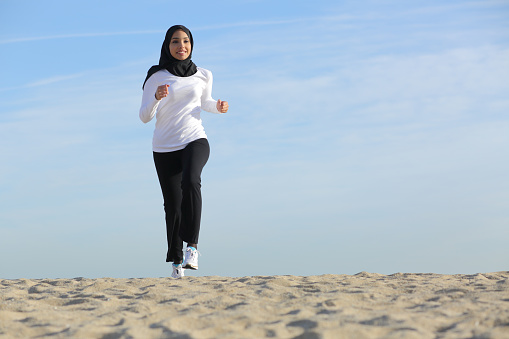 Front view of an arab saudi emirates woman running on the beach with the horizon in the background