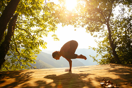 Young woman practicing yoga or doing just a stretching exercise next to the beautiful view on the canyon. She is wearing oriental trousers and a nude shirt. Taken by the Bosnian Croatian border, in the Dinara mountains, South Eastern Europe. A wooden terrace is made just next to the trees, lit by sun, a perfect place for meditation, exercise, yoga practice or just relaxation.
