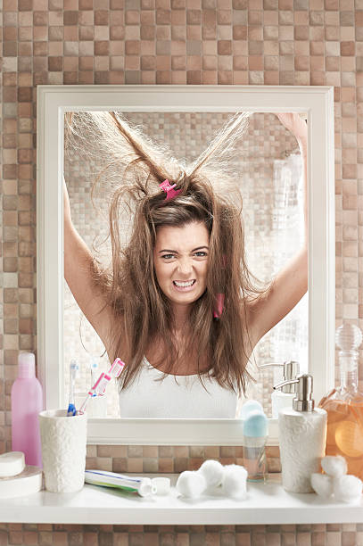 bad hair day a young woman stands in the bathroom looking in a mirror. She is trying everything to fix her hair but it doesn't seem to be working. angry hairstylist stock pictures, royalty-free photos & images