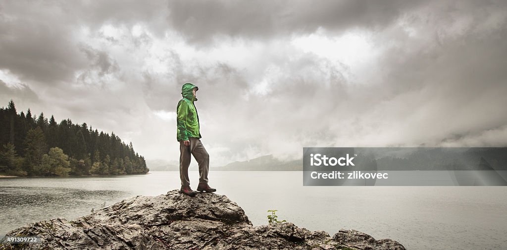 Man beside a mountain lake in rain Man standing on a rock beside a dramatic mountain lake after a hike in the rainy, gloomy day. Active lifestyle, outdoor activities, moods and emotions concept. Hiking Stock Photo