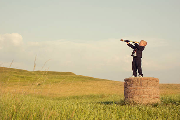 Young Boy Businessman Looking throught Telescope A young British boy in a business suit is standing on a pedestal looking through a hand-held telescope in the English countryside. He is looking for more business opportunities. telescope photos stock pictures, royalty-free photos & images
