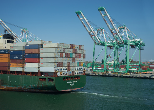 Container transportation is now the most popular way of cargo movement and a critical pillar of the global economy, Drone shot of a cargo ship sitting in dock in the Port of Long Beach, with crane booms lowering into position to unload the containers.