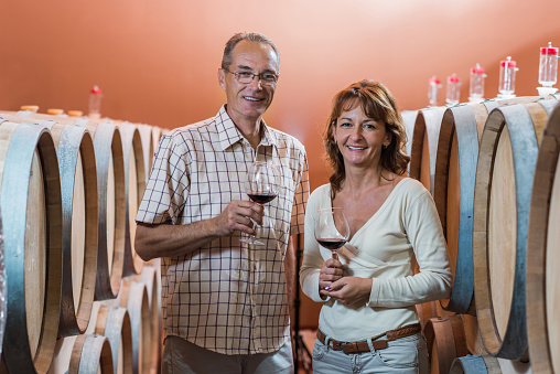 Mature couple standing in wine cask and holding a glass of red wine. They are looking at the camera.