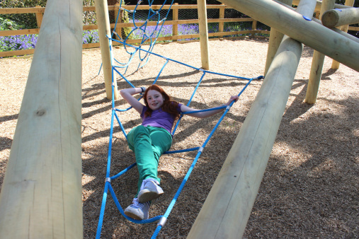 Photo showing a young girl with long red hair, dressed in a purple T-shirt and green jeans, playing in a woodland playground.  She is pictured lying on blue rope netting within a large climbing free made with wooden logs and posts, which appears quite natural and rustic.