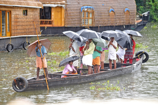 Kerala, India, February 19th 2013; A ferryman labours with his rudimentary boat through an unseasonal rainstorm as his passengers stand bedraggled under their umbrellas and await arrival at the opposite backwaters canal side as they pass traditional houseboats en route