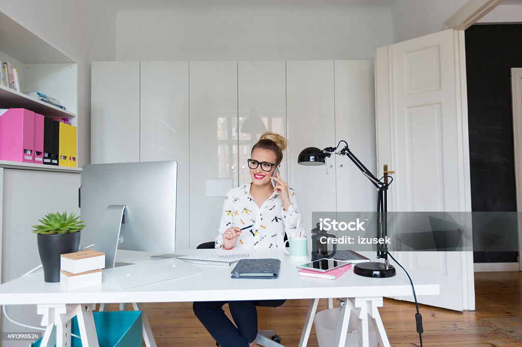 Woman working an office, talking on phone Front view of an attractive blonde woman sitting at the desk in an office and talking on the phone. Computer on the desk. 2015 Stock Photo