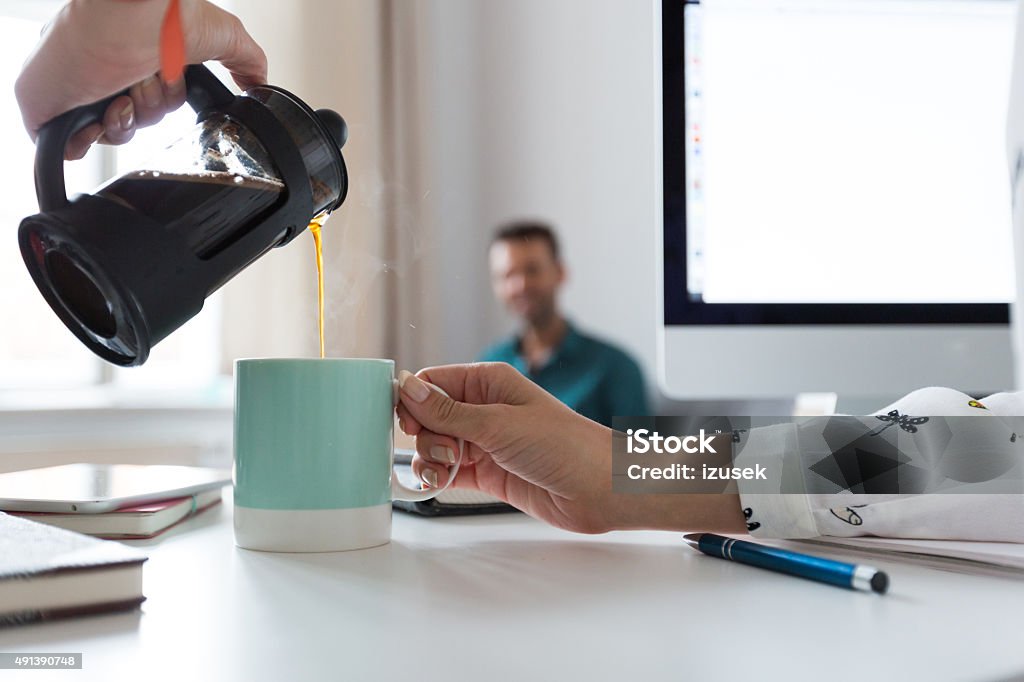 Woman pouring coffee into the cup in an office Woman pouring coffee from french press into the cup in an office. Close up of hands. Defocused man in the background. Office Stock Photo