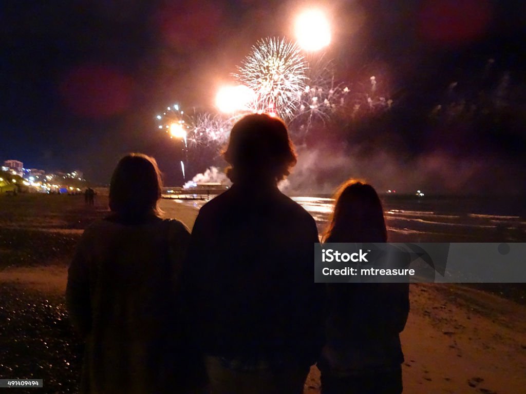 Image of family silhouettes watching seaside fireworks display on beach Photo showing the silhouettes of a family, watching a seaside fireworks display from the beach, with the sky lit up by the colourful pyrotechnics overhead. Firework Display Stock Photo