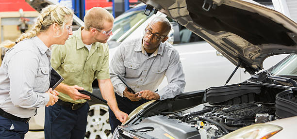 Team of multi-racial auto mechanics working on a car A team of three multi-ethnic auto mechanics in a repair shop, working on a car.  They are looking under the hood, holding digital tablets, discussing what needs to be repaired.  The African American man is in his 50s and the Caucasian man and woman are in their 20s. multiengine stock pictures, royalty-free photos & images