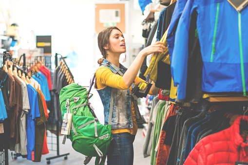 Female in sports and outdoor equipment store looking to buy some equipment. She is wearing green backpack and checking the wind jackets. Other items are visible in the store.