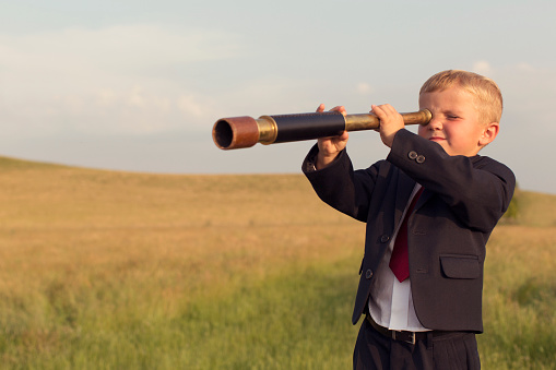 A young British boy in a business suit is standing on a pedestal looking through a hand-held telescope in the English countryside. He is looking for more business opportunities.
