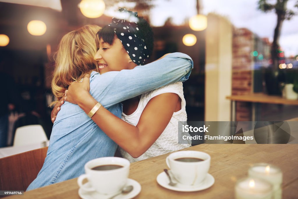 Two affectionate girl friends embracing Two multi ethnic affectionate girl friends embracing as they sit in a coffee shop enjoying a cup of coffee together Cafe Stock Photo