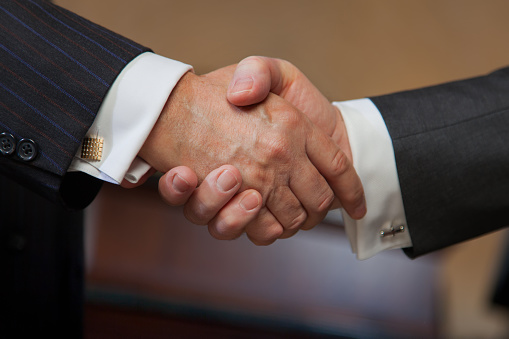 Two businessmen are shaking hands. They are in the meeting room of a tall office building.