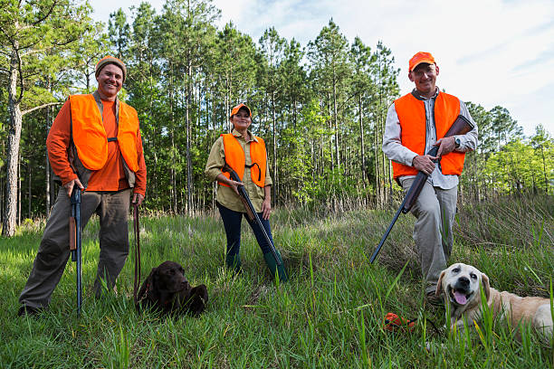 A group of three hunters, a woman and two men, with hunting dogs, standing in the tall grass by the woods, wearing orange safety vests and hats, smiling at the camera.  They are hunting birds or other game with shotguns.