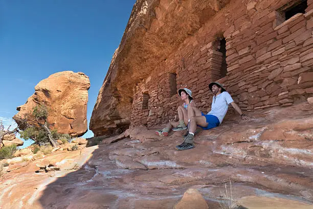Sitting near a massive balanced red rock boulder, a pair of trail runners/hikers, a man and woman, look out over the canyon country from the shade of a rock overhang housing extremely well preserved Ancestral Pueblo ruins that were once occupied around 1250 A.D. on the Cedar Mesa in Utah.