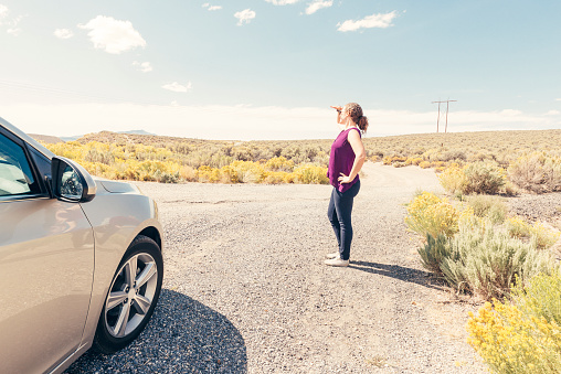 This is a horizontal, color, royalty free stock photograph of a empty dirt road off Highway 50 in rural Utah. The point of view is from the side of a car on the road less traveled.  A redhead in her 30s traveling by herself stops to look at the desert view while standing outside of her car. Flowering sagebrush grows all along the roadside. Photographed on a bright summer day with a Nikon D800 DSLR camera.