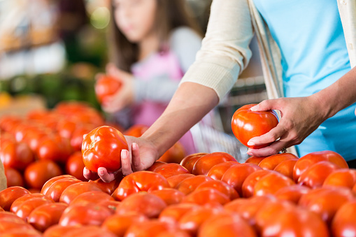 A woman is handling fresh tomatoes at a supermarket or local grocery store. Her daughter is also choosing vegetables in the background, adding depth and context to the image.