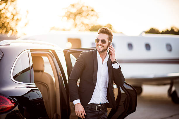 Man on the airport with phone in his hand Young well dressed man with sunglasses exiting from the back seat of the car and holding his smart phone. He looks like a famous musician or other celebrity. Private jet airplane is in the background. rich man stock pictures, royalty-free photos & images