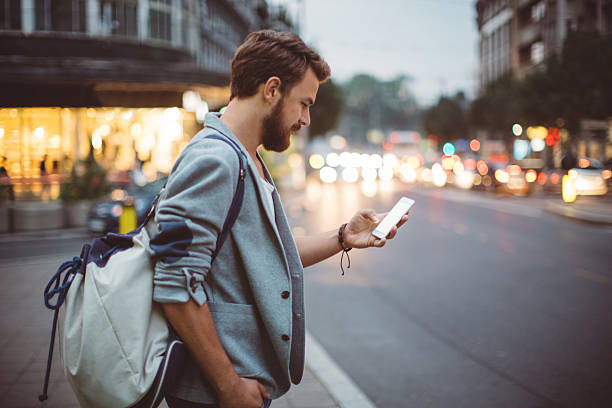 Young man on the streets of big city. Young man on the street of big city , waiting taxi, chacking his smartphone, for news or new messages. Or looking for map instructions. Carry backpack on one shoulder. Dusk time. Casual dressed. Street lights in background. mobile phone lights up stock pictures, royalty-free photos & images
