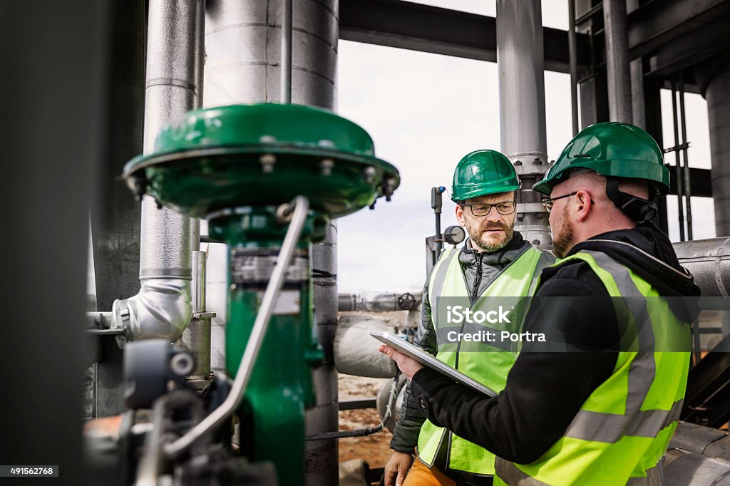 Male workers standing and discussing at factory Male worker talking to colleague in factory. They are discussing by machinery at industry. Professionals are wearing hardhats. Factory Stock Photo