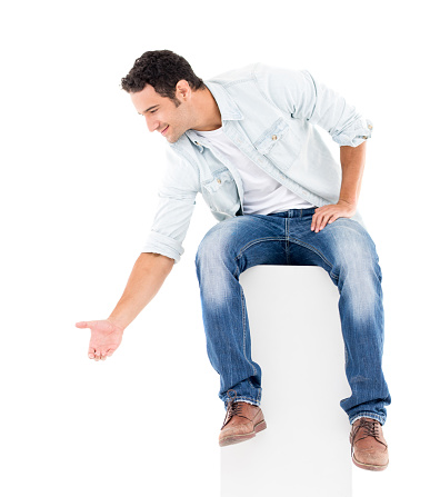 Casual man sitting on something and offering a helping hand - isolated over a white background