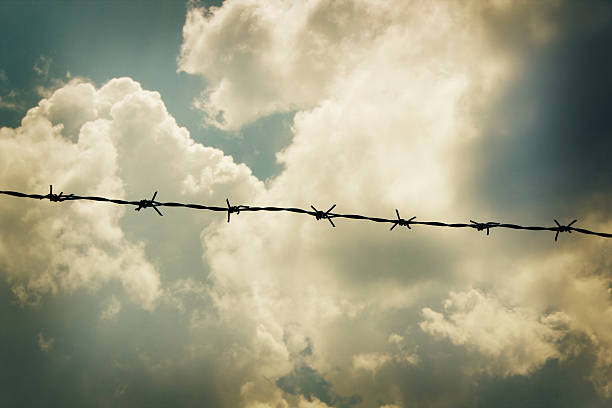 Fear and Hope, barbed wire against dramatically illuminated cloudy sky Fear and Hope, barbed wire against dramatically illuminated cloudy sky, metaphorical image. apartheid sign stock pictures, royalty-free photos & images