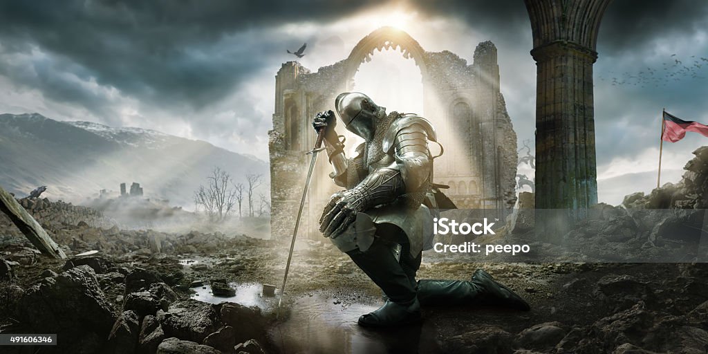 Medieval Knight Kneeling With Sword In Front of Building Ruin A Medieval knight wearing full suit of armour, boots and chainmail, kneeling as if in defeat or contemplation in preparation for battle. He rests on his sword in a puddle amongst rocks and rubble in front of a building ruin under a dramatic stormy evening sky with rays of sunlight. Knight - Person Stock Photo