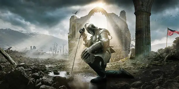 A Medieval knight wearing full suit of armour, boots and chainmail, kneeling as if in defeat or contemplation in preparation for battle. He rests on his sword in a puddle amongst rocks and rubble in front of a building ruin under a dramatic stormy evening sky with rays of sunlight.