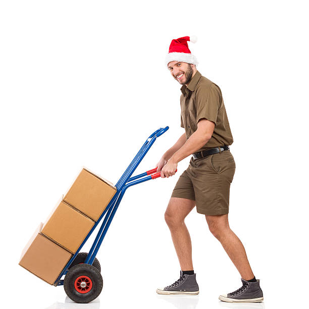 Christmas Messenger Delivering Packages Happy delivery man in santa claus hat pushing a delivery cart and looking at camera. Full length studio shot isolated on white. mover delivery person box hand truck stock pictures, royalty-free photos & images