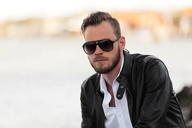 An attractive man in his 20s wearing a white shirt and black leather jacket with dark sunglasses, being by the water in a harbor. 